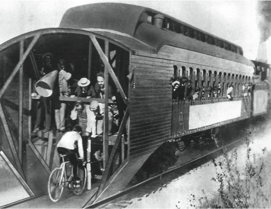 A black-and-white photograph of people on a train peeking out the windows at a man following the train on a bike.